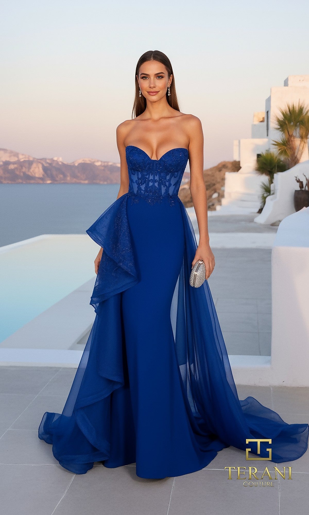 A woman in a royal Terani 261P6261 long formal dress stands poolside outdoors, holding a silver clutch, with white buildings and a scenic background.